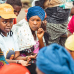 A group of Congolese women wearing colorful headscarves and clothing are sitting and engaging in conversation. Dr. Celestine Buyibuyi, in an Ipas orange cap and white shirt, is speaking to others who are attentively listening. The setting appears to be outdoors.