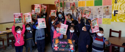 A group of children wearing face masks stand in a classroom holding up comic books. They appear to be raising awareness about childhood sexual violence in Bolivia, as they hold a poster that reads, 