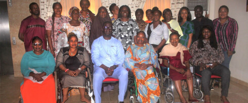 A diverse group of people, exemplifying disability inclusion, gather for a group photo indoors. Some use wheelchairs while others stand or sit. They are smiling and dressed in colorful attire, arranged in two rows against a backdrop of light-colored walls and curtains.