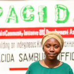Abdia Lalaikipia stands in front of a white signboard that reads "PACIDA Pastoralist Community Initiative and Development Assistance" and "PACIDA Sambuli Office" in black text. The person is wearing a green shirt and a beige headscarf, and is smiling at the camera.