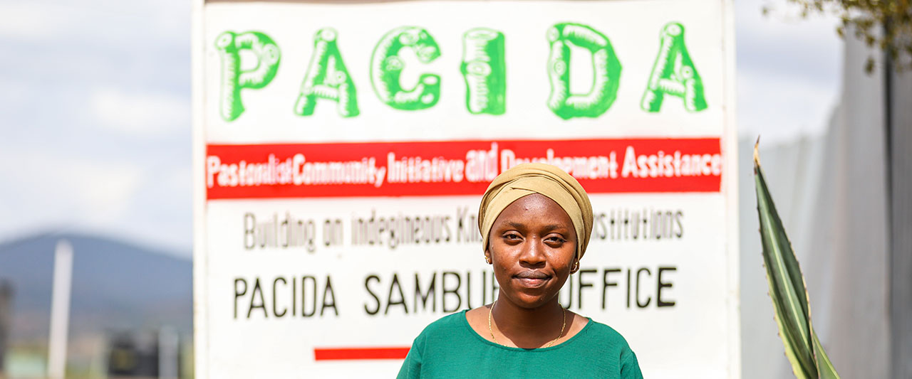 Abdia Lalaikipia stands in front of a white signboard that reads "PACIDA Pastoralist Community Initiative and Development Assistance" and "PACIDA Sambuli Office" in black text. The person is wearing a green shirt and a beige headscarf, and is smiling at the camera.