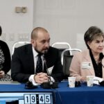 Two people sit at a conference table, a man and a woman, are speaking into microphones about child marriage, while the third person is focused on their phone. There is signage labeled "PETICIONARIO" on the table. Cups, microphones, and a clock displaying "19:45" are also visible.