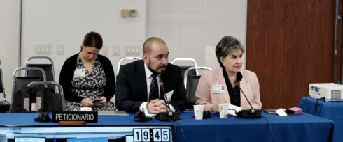 Two people sit at a conference table, a man and a woman, are speaking into microphones about child marriage, while the third person is focused on their phone. There is signage labeled 