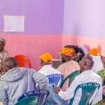A group of people, some wearing Ipas orange caps, sits in colorful plastic chairs. They are listening attentively to a person dressed in traditional attire seated in front of a banner about gender-based violence and a chalkboard. The room has purple and pink walls.
