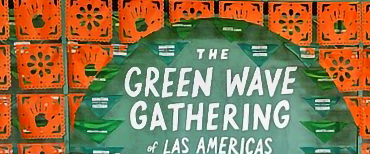 A sign reads: "The Green Wave Gathering of Las Americas," with a background of orange decorative paper cuttings featuring handprints and floral designs.