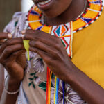 Close-up of a person wearing a bright yellow top and colorful beaded necklace, holding a yellow menstrual cup with both hands. The person is smiling, and adorned with beautifully patterned fabric on the shoulder. Trees are blurred in the background.