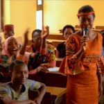A young woman in an orange dress stands speaking into a microphone in a Nigerian classroom full of smiling students who are raising their hands in joy reacting to her speech