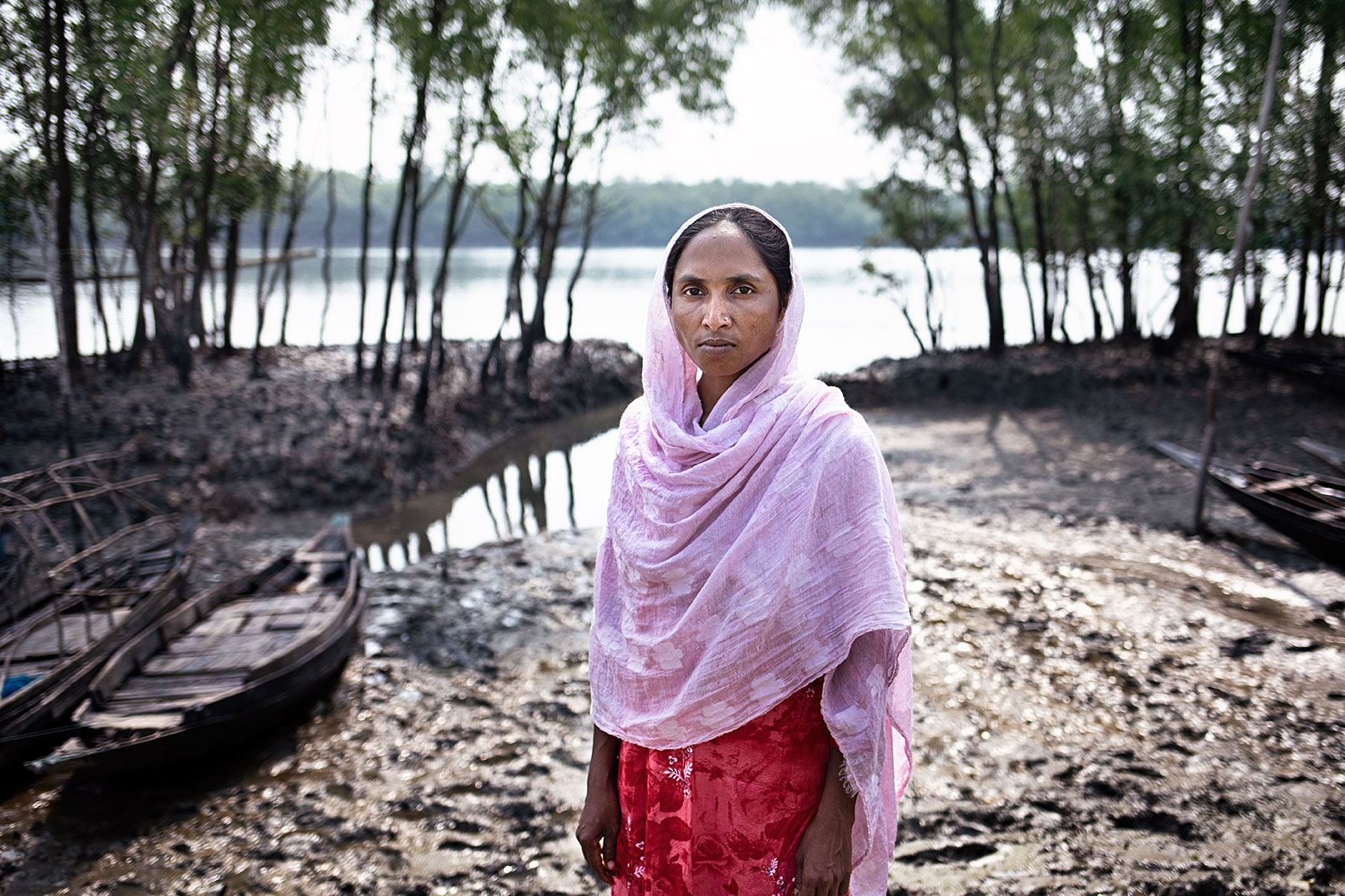 Selena Parvin stands on muddy ground near a body of water, surrounded by trees and small wooden boats. She is wearing a light pink headscarf and a red outfit. The background is a mix of natural elements, indicating a rural or natural setting, likely in a coastal area.
