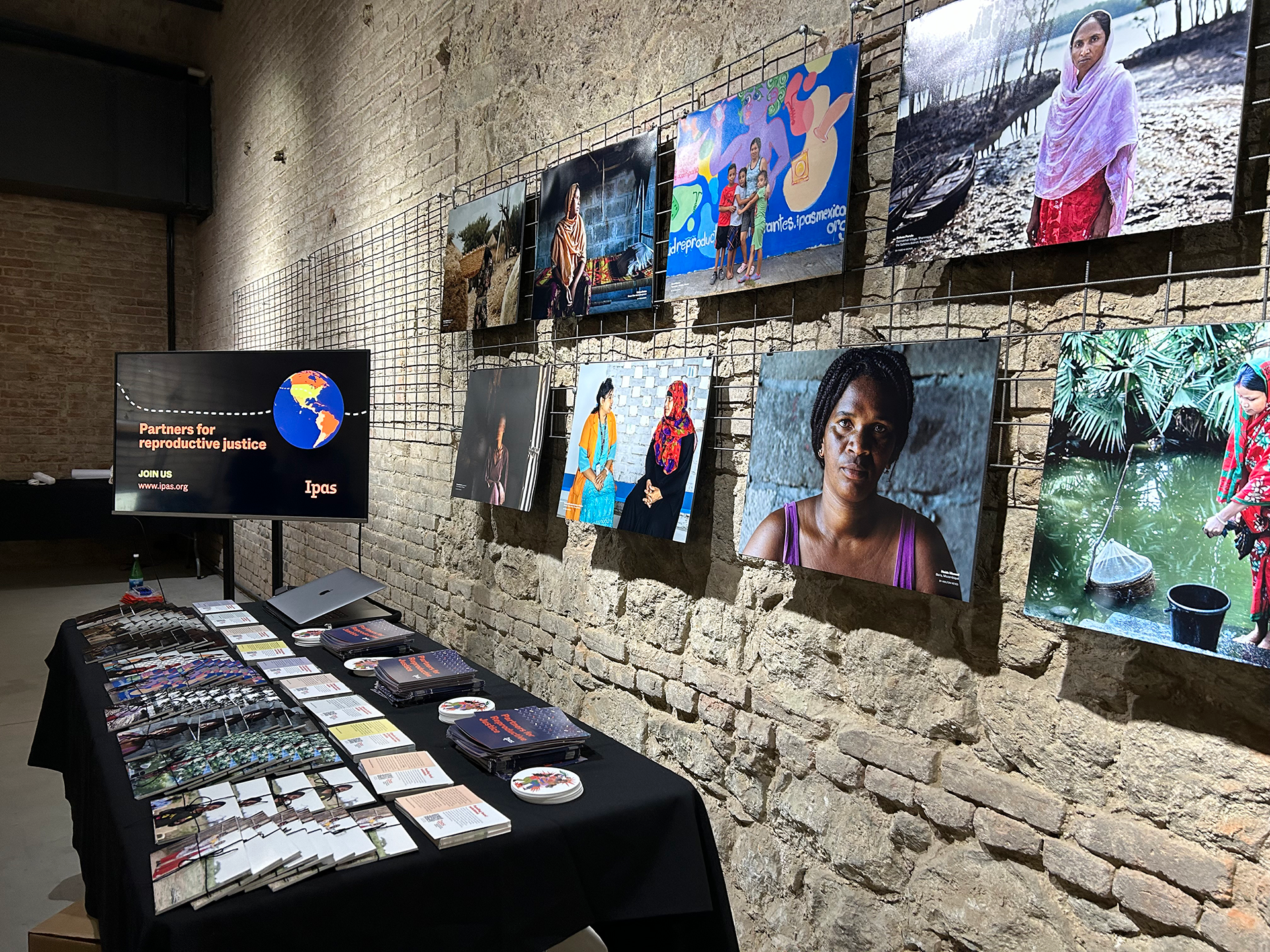 A dimly lit gallery showcases a series of photographs of diverse women on a textured brick wall. Below, a table is arranged with pamphlets, booklets, and promotional materials. A screen on the left reads "Partners for reproductive justice" with the Ipas logo.