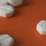Misoprostol pills image reveals a scattered display of white, hexagonal tablets on an orange surface, some with visible imprints. The tablets are arranged in small clusters, hinting at the presence of multiple individual doses.