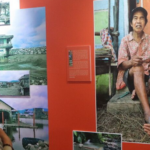 Two women, one in a beige hijab and the other in a red hijab, engage in conversation in front of a large photographic exhibition. The exhibits display images, primarily of rural scenes and a prominent portrait of an elderly woman sitting on a step.