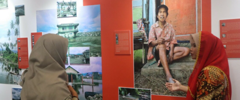 Two women, one in a beige hijab and the other in a red hijab, engage in conversation in front of a large photographic exhibition. The exhibits display images, primarily of rural scenes and a prominent portrait of an elderly woman sitting on a step.