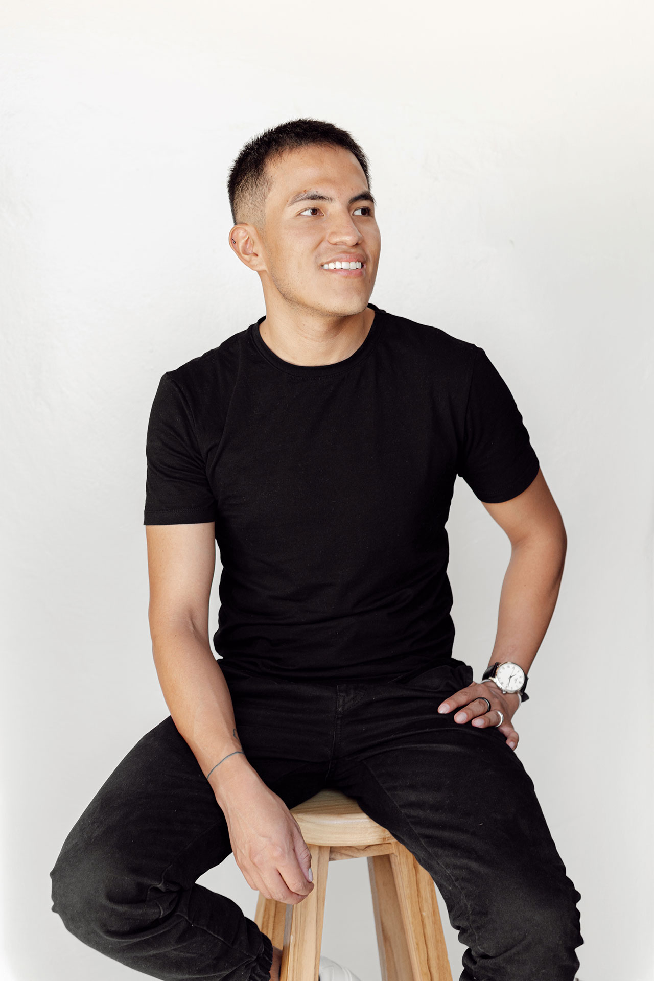 A person with short dark hair sits on a wooden stool against a plain white background. They are wearing a black t-shirt, black jeans, and a watch on their left wrist, and they are smiling while looking slightly to the right.