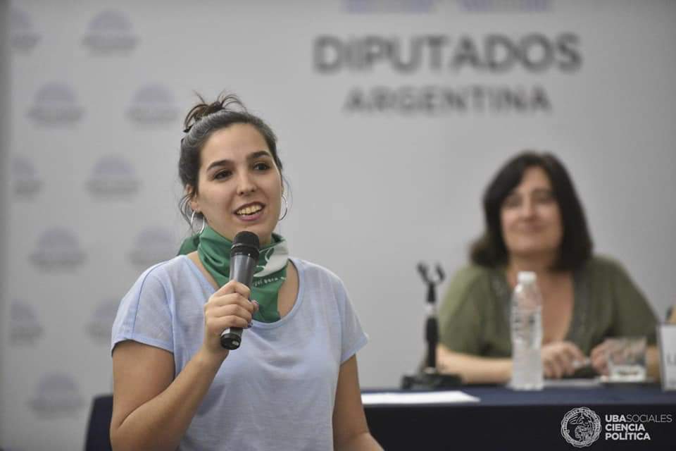 A woman holds a microphone and speaks at an event. She wears a green bandana around her neck and stands in front of a banner that reads, "Diputados Argentina." Another woman sits at a table in the background with a microphone and a water bottle.