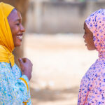 Two women, Maryam Umar (left) and Rahama Hassan (right) stand facing each other, smiling and chatting. Umar wears a yellow headscarf and blue patterned dress, with a backpack on her shoulder. Hassan wears a pink headscarf with dark blue spots and a light purple patterned dress. A tree is in the background.