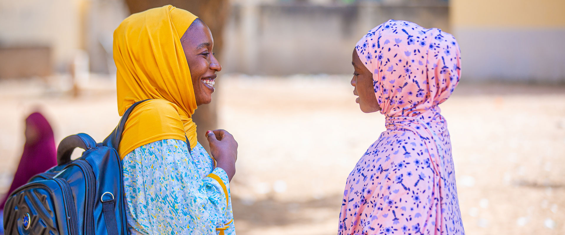 Two women, Maryam Umar (left) and Rahama Hassan (right) stand facing each other, smiling and chatting. Umar wears a yellow headscarf and blue patterned dress, with a backpack on her shoulder. Hassan wears a pink headscarf with dark blue spots and a light purple patterned dress. A tree is in the background.