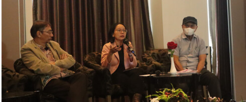 Three people are seated on stage during a panel discussion. The person in the middle is speaking into a microphone, while the other two listen attentively. A table in front of them holds a single red flower in a vase and some papers. A dark curtain hangs in the background.