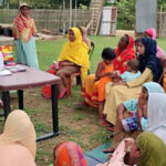 A group of women, many in colorful traditional clothing and headscarves, sit outdoors on chairs and the ground around a table. Two healthcare workers in white coats stand and give a presentation, displaying educational materials to the attendees.