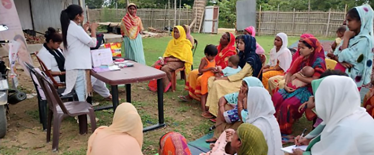 A group of women, many in colorful traditional clothing and headscarves, sit outdoors on chairs and the ground around a table. Two healthcare workers in white coats stand and give a presentation, displaying educational materials to the attendees.