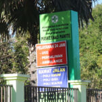 A signboard for a healthcare facility stands amidst greenery and trees. The green board has various sections with text indicating services like emergency department (UGD), maternity, ambulance, and outpatient clinics including general, dental, and KIA services.