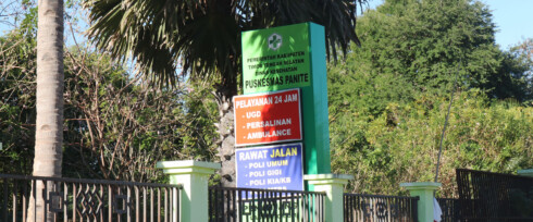 A signboard for a healthcare facility stands amidst greenery and trees. The green board has various sections with text indicating services like emergency department (UGD), maternity, ambulance, and outpatient clinics including general, dental, and KIA services.