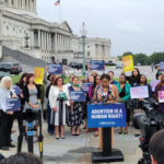 A group of people is gathered outside a government building holding signs supporting abortion rights. A woman is speaking at a podium with a sign reading, "Abortion Is a Human Right!" Reporters and cameras are in the foreground capturing the event.