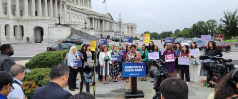 A group of people is gathered outside a government building holding signs supporting abortion rights. A woman is speaking at a podium with a sign reading, 