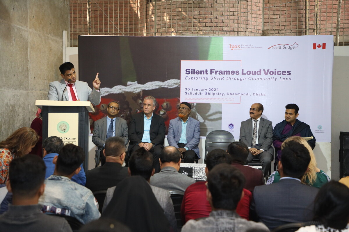 A man in a red tie speaks at a podium in front of an audience. Behind him, a banner reads "Silent Frames Loud Voices: Exploring SRHR through Community Lens," with logos from Ipsos and Global Affairs Canada. Five seated men are on stage, attentively listening.