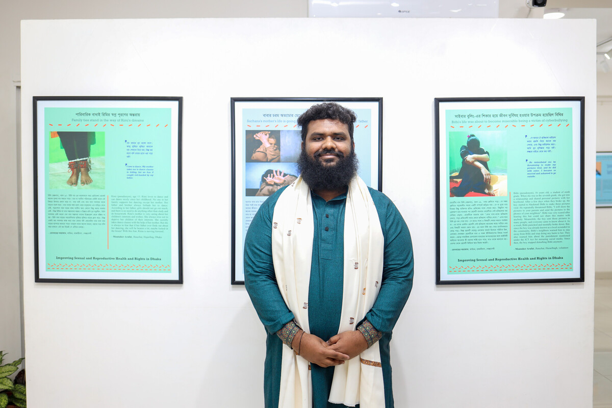 A man with a beard and dark hair, wearing a teal kurta and white shawl, stands in front of a wall displaying three framed informational posters. The posters contain text and images, possibly part of an exhibition. The room appears to be a gallery or display area.