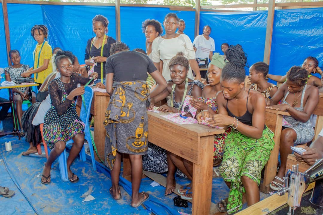 A group of women and young girls are gathered in a vibrant, makeshift classroom. They are all engaged in sewing activities, using colorful fabrics. The classroom has wooden desks and blue tarp walls, creating a lively and collaborative atmosphere for learning.