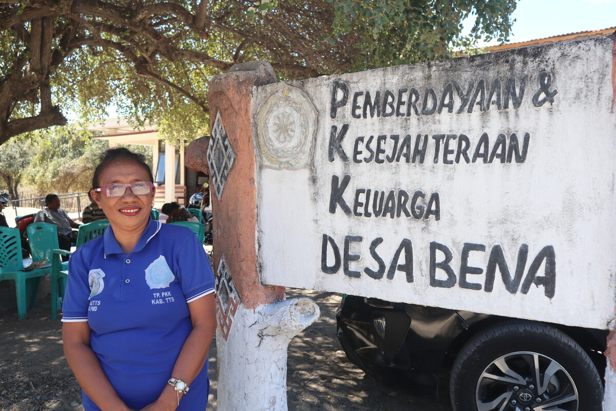 A woman wearing a blue polo shirt and glasses stands smiling beside a large weathered sign that reads, "Pemberdayaan & Kesejahteraan Keluarga Desa Bena." Trees and several people sitting in green chairs are visible in the background.