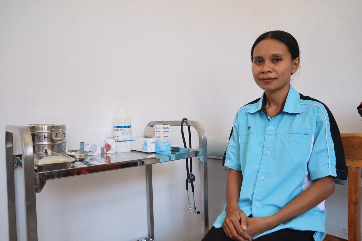 A healthcare worker wearing a light blue uniform sits beside a medical cart containing various supplies, such as a stethoscope, alcohol bottle, and medical boxes, against a plain white wall background.