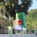 A signboard in front of a health clinic (Puskesmas Painte) in an outdoor setting with trees in the background. The sign lists services available: 24-hour emergency services, obstetrics, ambulance, general outpatient care, dental care, and basic health.