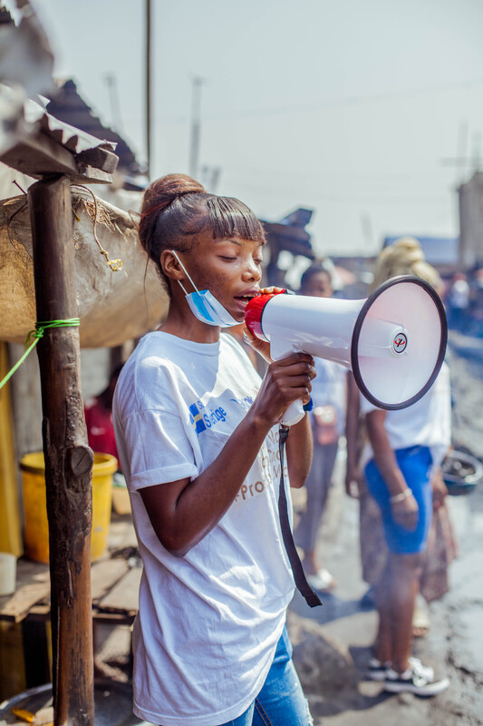 A young woman with braids, partially wearing a face mask, holds a megaphone and speaks while standing in an outdoor setting. She is wearing a white shirt, and there are blurred figures and structures in the background, indicating a community environment.