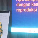 Nisa Anisa, wearing glasses and a plaid shirt is standing and speaking into a microphone, gesturing with one hand. Behind them is a presentation slide in Indonesian and a whiteboard with handwritten notes. The background has vertical dark panels.