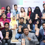 A group of people, some seated and some standing, hold up awards and smile at the camera. They are dressed in a mix of formal and traditional attire. A few banners in the background display logos and text, and the flags of Canada and Bangladesh are partially visible.