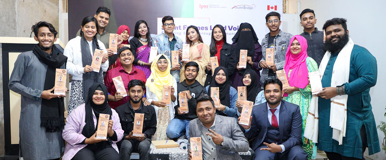 A group of people, some seated and some standing, hold up awards and smile at the camera. They are dressed in a mix of formal and traditional attire. A few banners in the background display logos and text, and the flags of Canada and Bangladesh are partially visible.