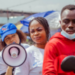 A group of people outdoors, one woman holds a megaphone, wearing a blue cap and white shirt. Another woman stands beside her, and a man in a red hoodie is in the foreground. Market stalls and umbrellas are visible in the background.