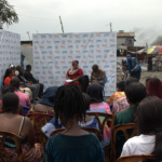 A community gathering is taking place outdoors. Numerous people are seated and facing a makeshift stage where individuals are speaking. The backdrop displays various logos. The setting appears to be in an underdeveloped area with visible structures and debris.