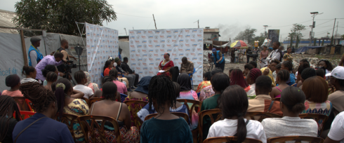 A community gathering is taking place outdoors. Numerous people are seated and facing a makeshift stage where individuals are speaking. The backdrop displays various logos. The setting appears to be in an underdeveloped area with visible structures and debris.