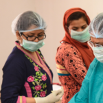 Four healthcare workers wearing surgical caps, face masks, and gloves are engaged in a medical procedure or training session. Three are dressed in colorful attire, while one wears a green medical gown. They are in a well-lit room with medical supplies.