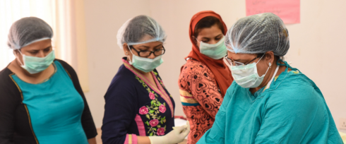 Four healthcare workers wearing surgical caps, face masks, and gloves are engaged in a medical procedure or training session. Three are dressed in colorful attire, while one wears a green medical gown. They are in a well-lit room with medical supplies.