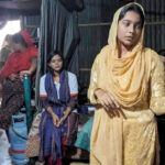 A youth volunteer from Bangladesh in traditional clothing presents to a group in a dimly lit room. She gestures towards a poster with text and illustrations while several women sit and listen attentively. Various household items are visible in the background.