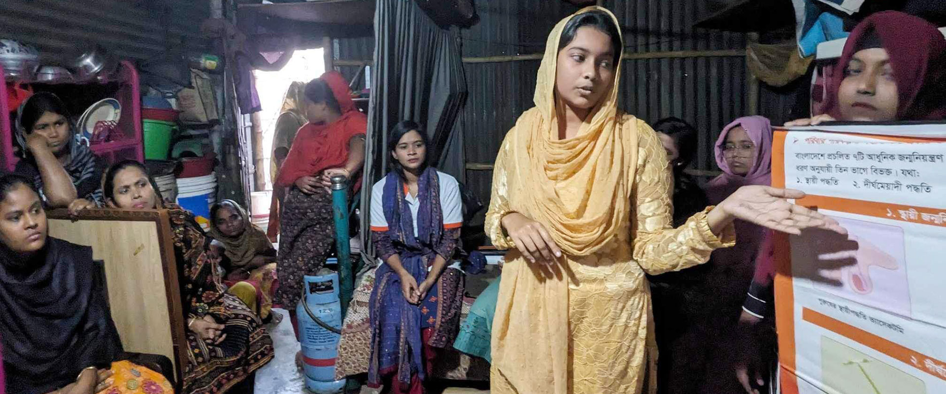 A youth volunteer from Bangladesh in traditional clothing presents to a group in a dimly lit room. She gestures towards a poster with text and illustrations while several women sit and listen attentively. Various household items are visible in the background.