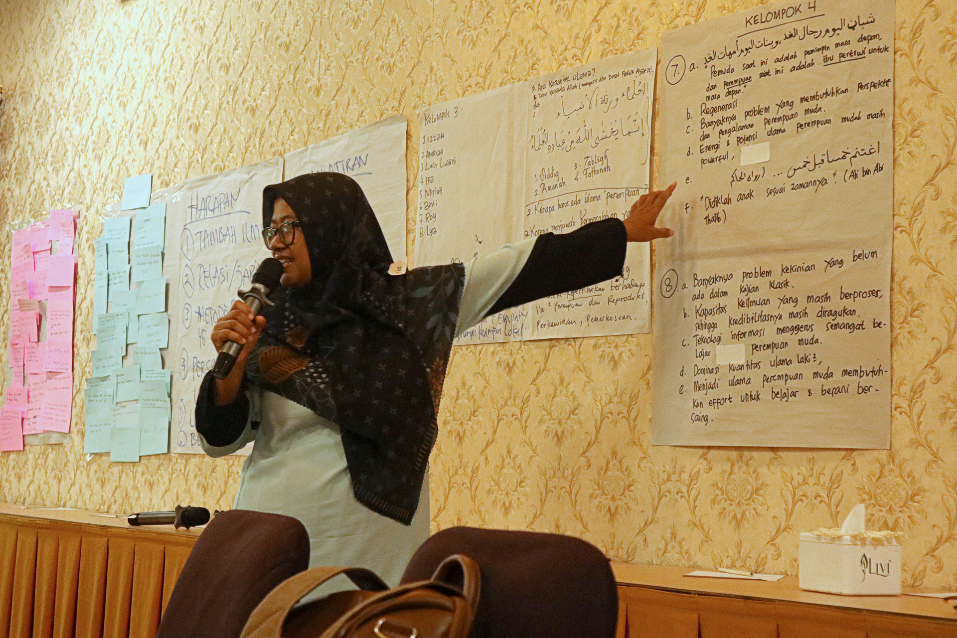 A woman wearing a hijab holds a microphone and points to a large piece of paper with writing on a wall. The room has several papers with notes and lists displayed, and she appears to be leading a presentation or workshop.