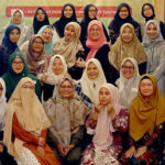 A group photo of 39 women and one child, many wearing colorful hijabs, posed indoors. In the background, there's a red banner and a whiteboard. The setting appears to be a social or educational event.