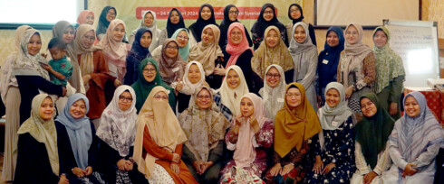 A group photo of 39 women and one child, many wearing colorful hijabs, posed indoors. In the background, there's a red banner and a whiteboard. The setting appears to be a social or educational event.