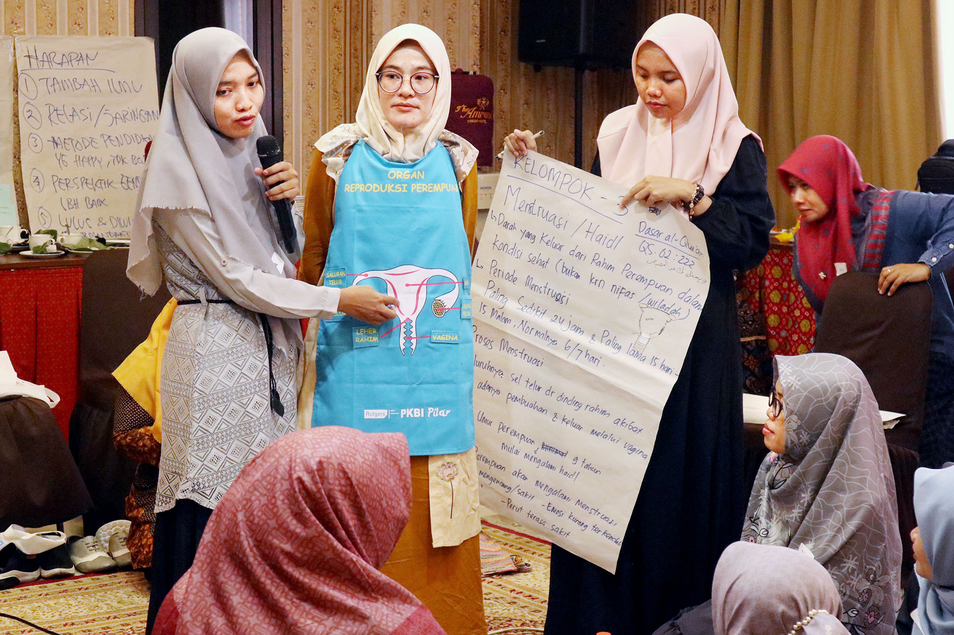 Three women in hijabs stand in a group discussion. One wears an apron with a uterus diagram, while another holds a large paper with notes. Participants sit around them, engaging in the presentation.