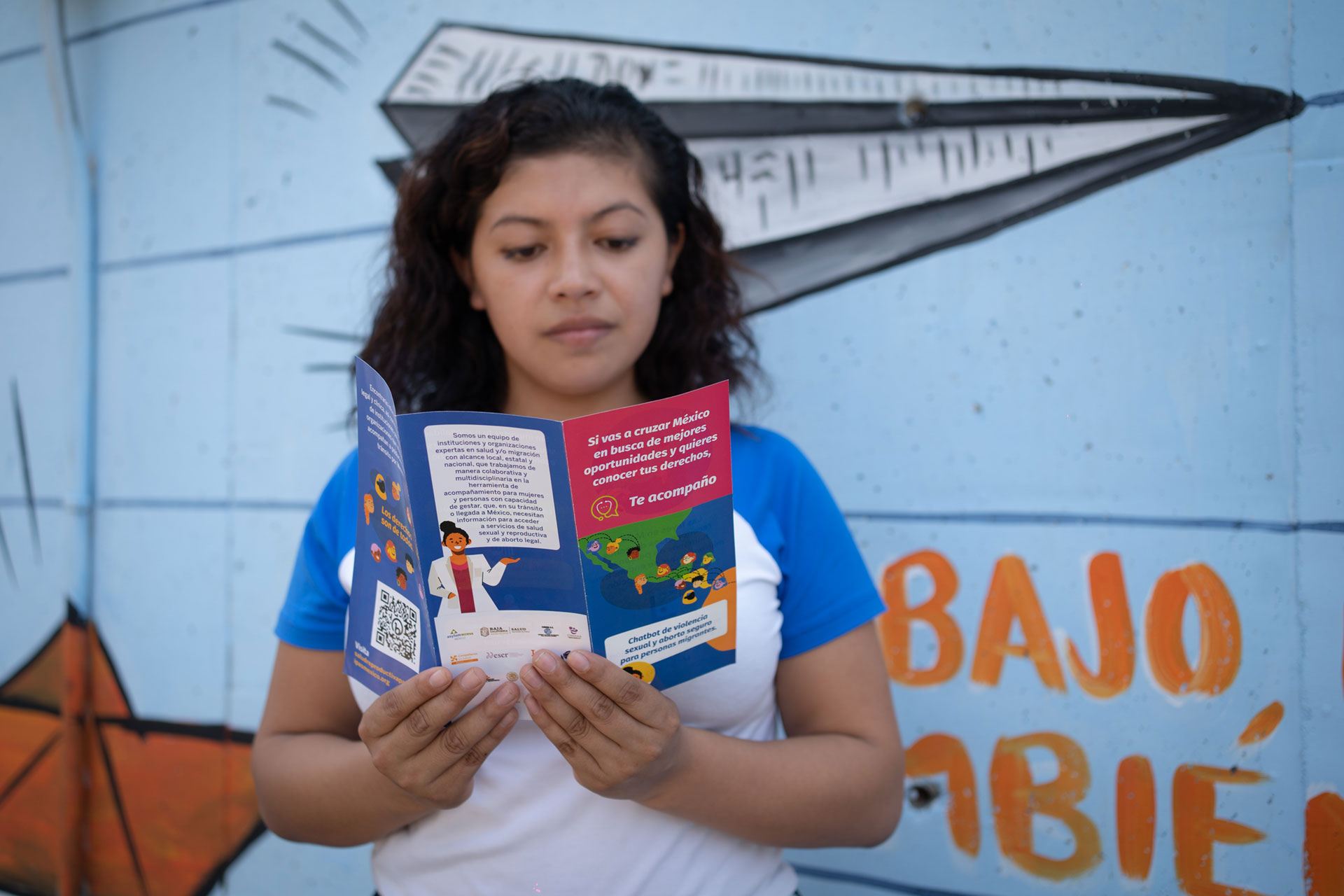 Elizabeth Martinez has curly hair, wearing a blue and white shirt, reads a colorful Te Acompaño chatbot brochure. The background features a mural with a paper airplane and text in Spanish. The scene is outdoors.