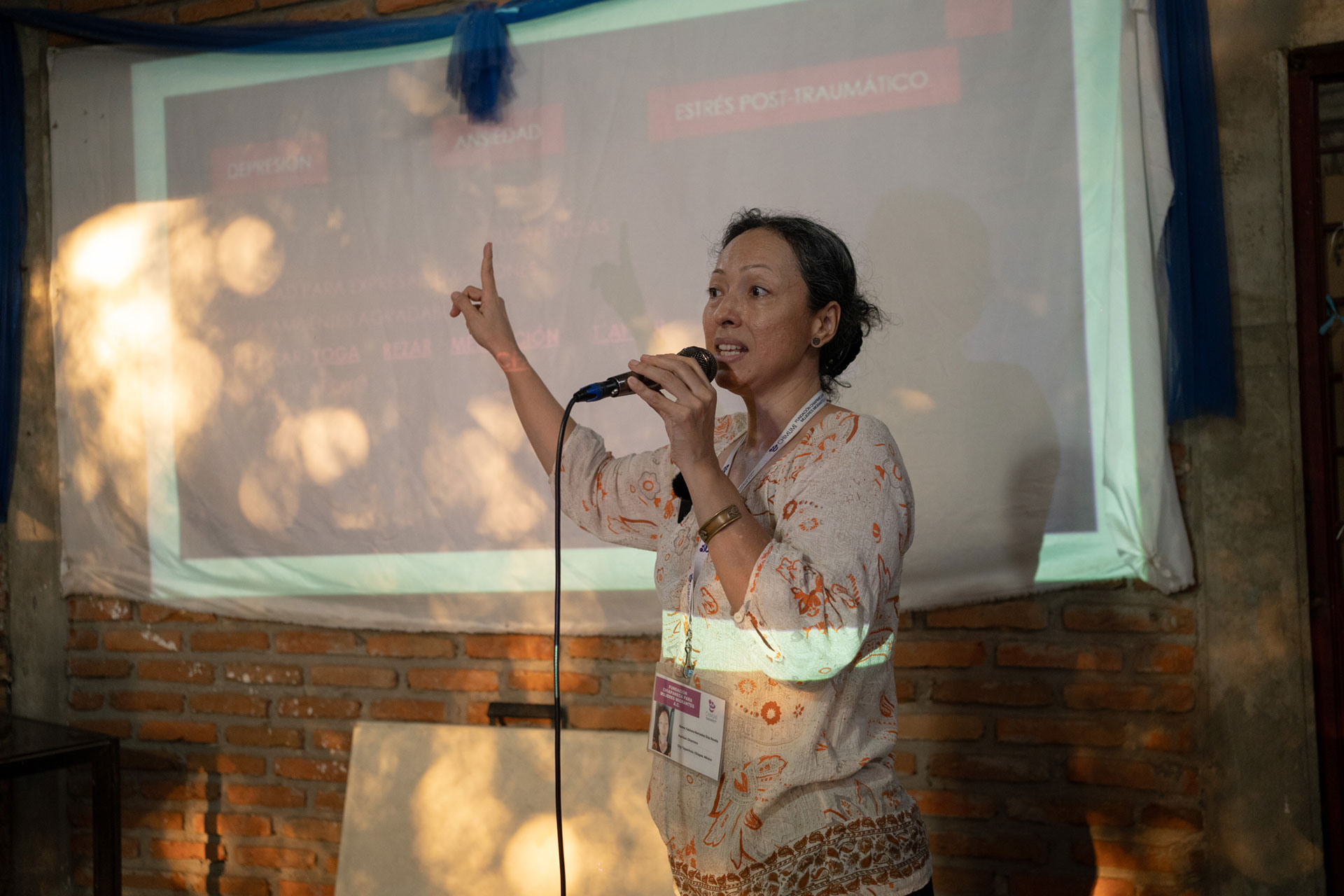 Fabiola Diaz Rovelo stands in front of a projected presentation, speaking into a microphone and gesturing with one hand. The presentation appears to be about "estrés post-traumático." The setting is indoors with brick walls.